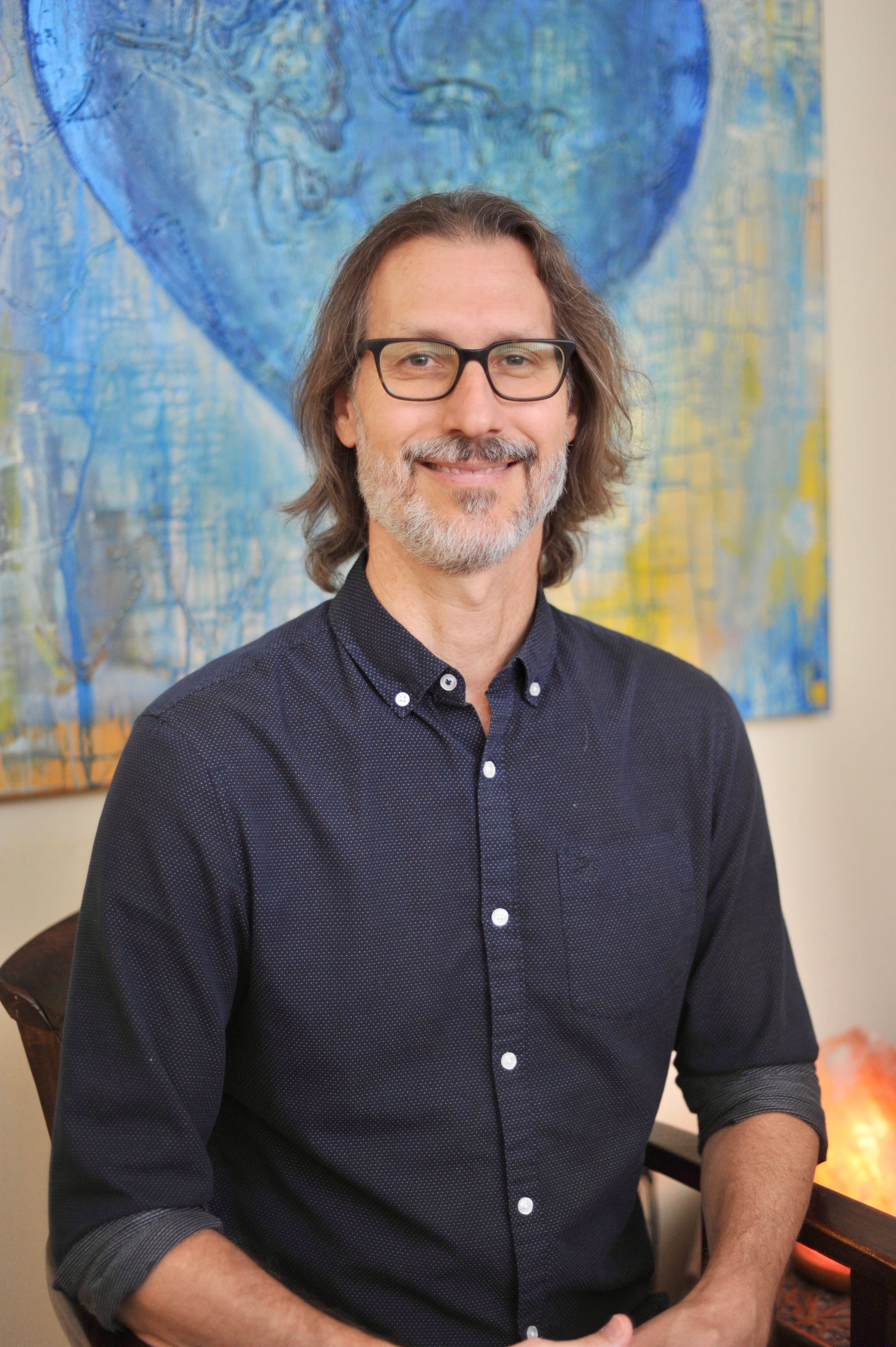 A Miami Beach Acupuncturist with glasses, a graying beard, and shoulder-length brown hair, wearing a dark navy blue button-down shirt, standing in front of a blue and yellow abstract painting.